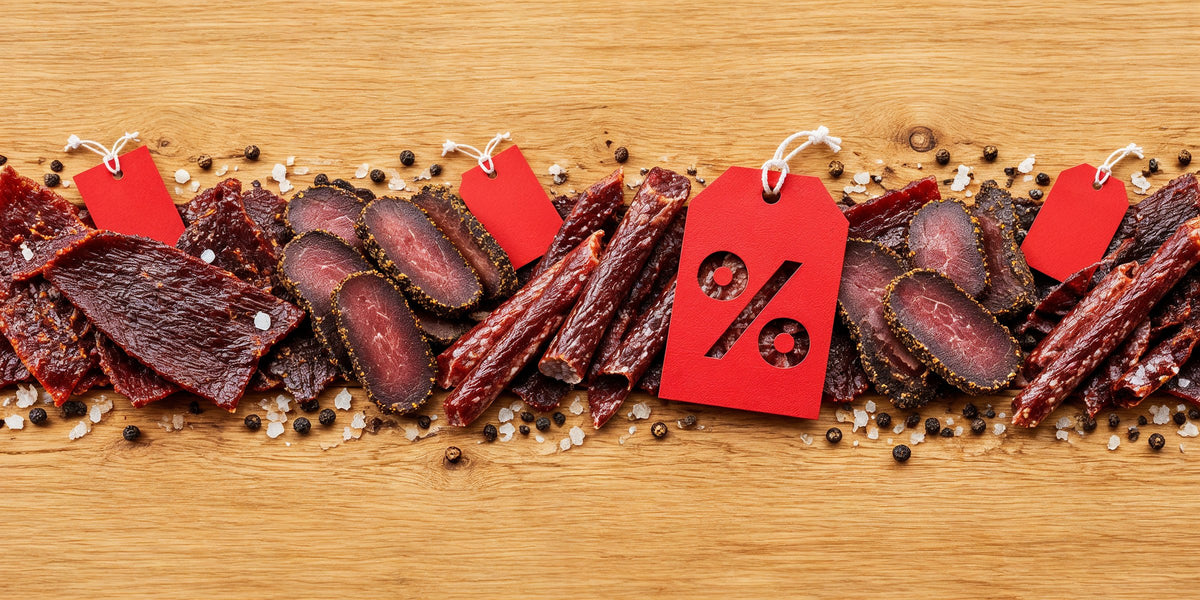 Jerky and meat snacks on sale: beef jerky, biltong and dried sausage sticks on a wooden table with red price tags and a percent tag