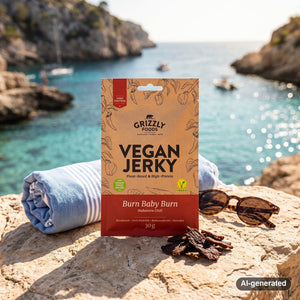 Grizzly Foods Vegan Jerky Burn Baby Burn on a rock by a Mediterranean cove, with a towel and sunglasses, AI-generated image